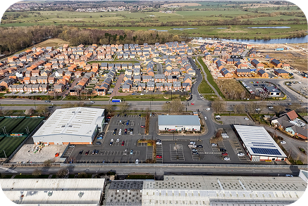 Aerial view of a suburban residential area with rows of houses, green spaces, parking lots, sports fields, and several commercial buildings, bordered by roads and open fields in the background.