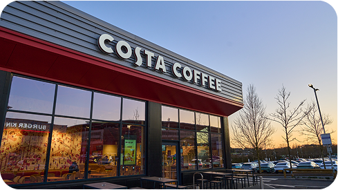 Costa Coffee shop exterior at sunset, with large windows reflecting the sky and parked cars in the background. The red and white Costa Coffee sign is prominently displayed above the entrance.
