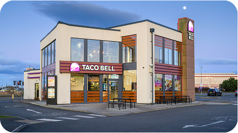 A modern Taco Bell restaurant with large windows and outdoor seating is shown at dusk, with the sky turning blue and a few cars parked nearby. The Taco Bell logo is displayed prominently on the building.