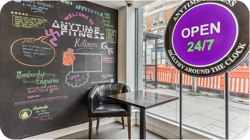 A black chalkboard wall with colorful fitness-themed drawings and messages is beside a glass window displaying a large purple OPEN 24/7 sign. A black chair and a small table are placed by the window.