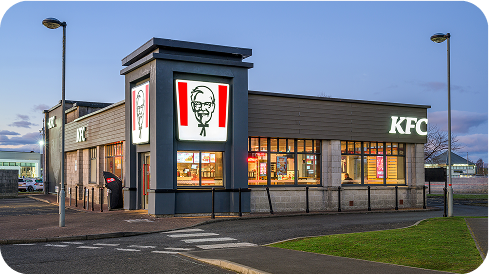 A modern KFC restaurant with large windows and illuminated KFC signage, including the Colonel Sanders logo, at dusk with streetlights and an empty parking area in front.