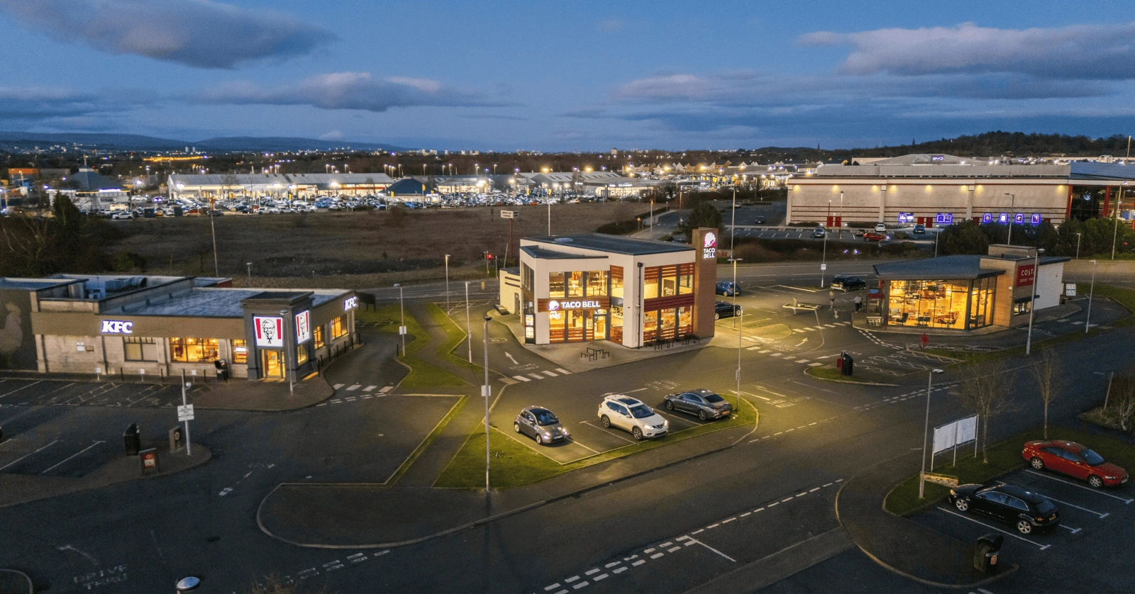 Aerial view of a commercial area at dusk with KFC, Pizza Hut, and Burger King restaurants, several parked cars, streetlights, and a large retail parking lot in the background.
