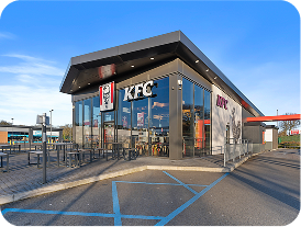 A modern KFC restaurant with glass walls, outdoor seating, and a drive-thru, set under a clear blue sky. Handicapped parking spaces are visible in the foreground.