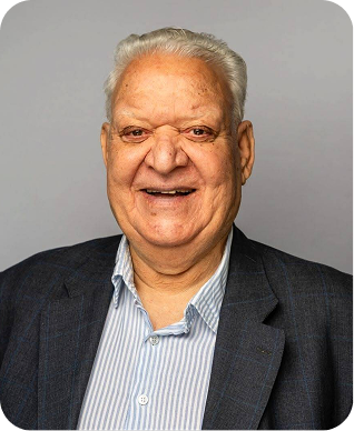 An elderly man with white hair and a big smile is wearing a navy blazer over a light blue striped shirt, posing in front of a plain gray background.