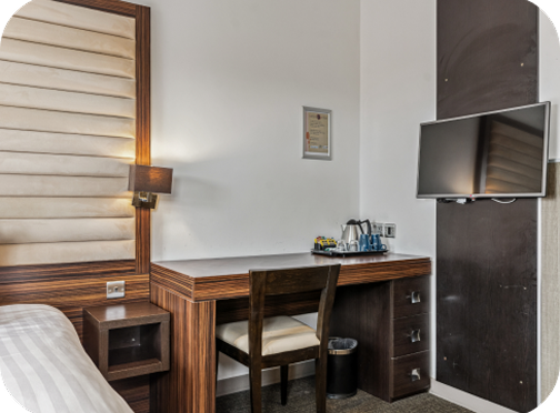 A modern hotel room workspace with a wooden desk, chair, tea and coffee set, wall-mounted TV, lamp, and part of a bed visible on the left. The decor features neutral tones and padded wall panels.