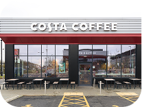 Exterior view of a Costa Coffee shop with large glass windows, outdoor seating, and a red and white sign above the entrance. A parking lot with a reserved space is in front of the café.