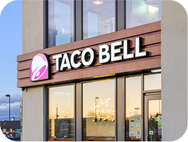The exterior of a Taco Bell restaurant with a large Taco Bell sign and logo above the entrance. The windows reflect the outdoor scene and evening sky.