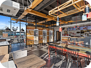Modern fast-food restaurant interior with self-service kiosks, wooden tables and chairs, pendant lights, and a counter in the background. Large windows let in natural light, and menu boards are visible above the counter.