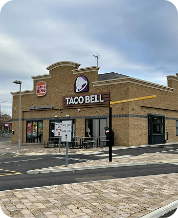 A Taco Bell restaurant with a Burger King sign on the building, showing drive-thru lanes, an outdoor menu board, and a few tables outside under a cloudy sky.