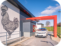 A white car in a fast-food drive-thru lane waits at the order window; a large rooster mural decorates the buildings exterior wall on a sunny day.