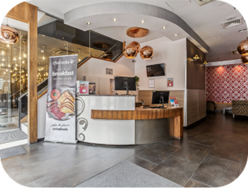 Modern hotel reception area with a curved front desk, computer monitors, copper pendant lights, a breakfast promotion sign, and a seating area with patterned wallpaper in the background.