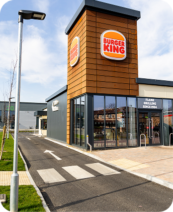 A Burger King restaurant with large signs, glass windows, and a drive-thru lane. The sky is partly cloudy and there is a pedestrian crossing near the entrance.