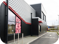 A modern drive-thru restaurant with black metal siding, large glass window featuring pink artwork, and a drive-thru lane with arrows on the pavement.
