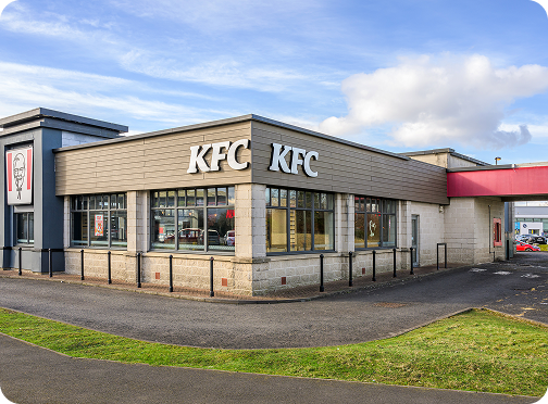 A KFC restaurant with large windows and the KFC logo on the building, surrounded by a paved driveway and a small grassy area under a partly cloudy sky.