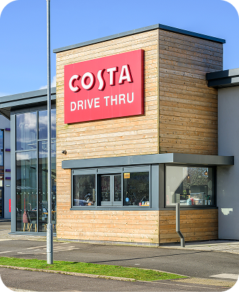 A Costa Coffee drive-thru building with a large red sign reading Costa Drive Thru above the service window, surrounded by modern wood and glass architecture on a sunny day.