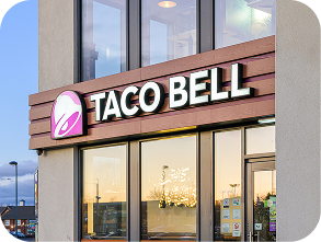 A Taco Bell restaurant exterior with a large sign displaying the Taco Bell logo and name above the entrance. The building reflects the sunset in its windows.