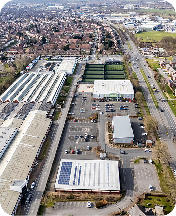 Aerial view of an industrial and commercial area with several large buildings, parking lots, and solar panels on one roof, adjacent to a main road and residential neighborhood in the background.