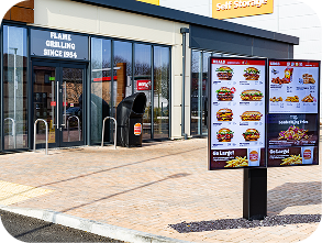 A fast-food restaurant exterior with glass doors and a drive-thru menu board displaying images of burgers, fries, and drinks. A sign above the door reads, “Flame Grilling Since 1954.”.