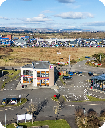 Aerial view of a fast food restaurant with drive-thru, parking spaces, and cars near a road, with fields and a large shopping complex in the background under a partly cloudy sky.