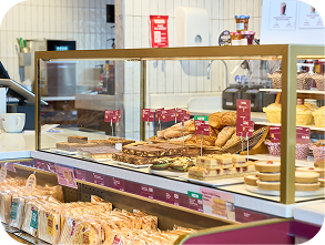 A bakery counter displays various pastries, cakes, and bread behind a glass case, with packaged baked goods on shelves below and a coffee cup on the counter.