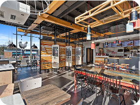 Modern fast-food restaurant interior with large windows, self-service kiosks, wooden tables and chairs, colorful pendant lights, and an open, well-lit dining area.