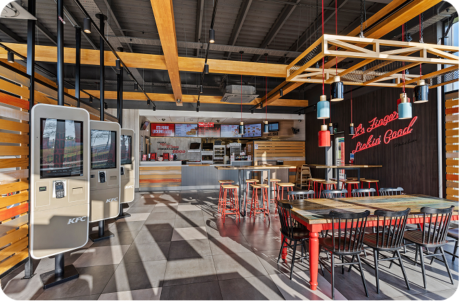 Modern KFC restaurant interior with self-order kiosks, wooden tables and chairs, pendant lights, and a counter in the background. A neon sign on the wall reads “It’s Finger Lickin’ Good.”.