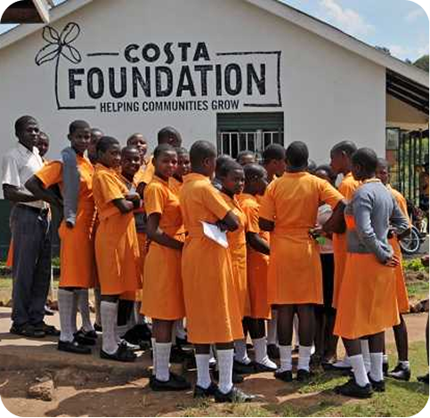 A group of students in orange uniforms stand outside a building with a sign reading Costa Foundation: Helping Communities Grow. An adult stands nearby, and the scene is bright and lively.