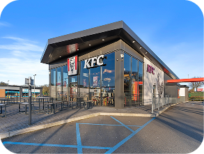 A modern KFC restaurant with large glass windows, outdoor seating, and a drive-thru lane. The KFC logo is visible above the entrance. The sky is clear and the parking lot includes accessible parking spaces.