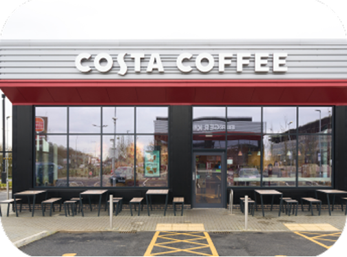 Front view of a Costa Coffee shop with large glass windows, outdoor seating, and a red and white sign. There are several tables and chairs outside, and a parking area with accessible spaces in front.