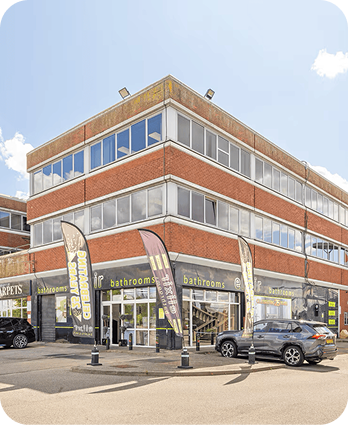 Three-story commercial building with large windows and red brick exterior. Ground floor features bathrooms and carpets stores, with flags promoting the businesses. Cars are parked in front on a sunny day.