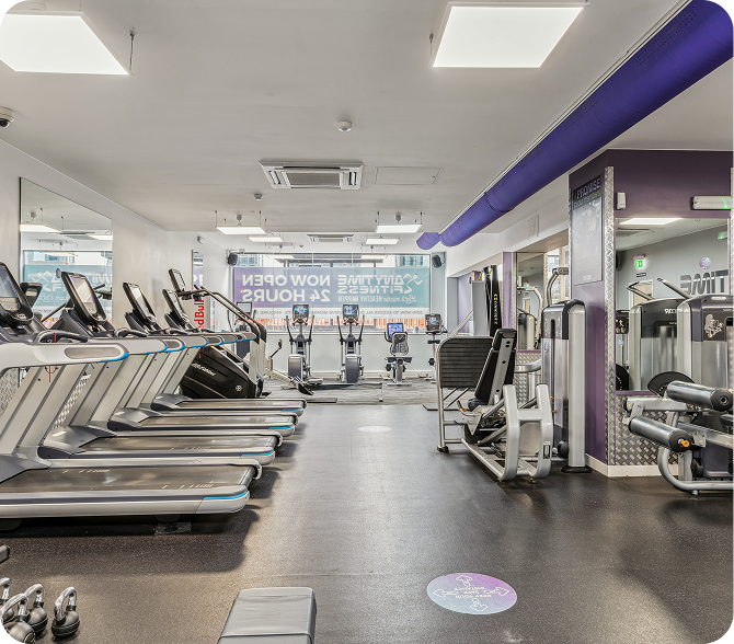 A modern gym interior with several treadmills on the left, various exercise machines on the right, and bright lighting. The room has mirrors and signage indicating it is open 24 hours.