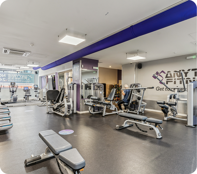 A modern gym with various exercise machines, benches, and weights. One person is using a weight machine. The space is clean, well-lit, and has large windows and purple accents.