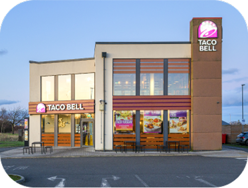 A Taco Bell restaurant with large windows, outdoor seating, and the Taco Bell logo displayed on the building and sign, seen in daylight with a clear sky.