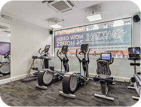 A small gym with exercise machines, including ellipticals and a recumbent bike, facing a large window. A sign outside reads Anytime Fitness Now Open 24 Hours. The room is well-lit with gray carpet flooring.
