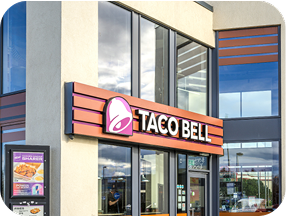 Exterior view of a Taco Bell restaurant with large windows, the Taco Bell logo and sign above the entrance, and a drive-thru menu board visible on the left side.
