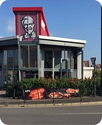 A KFC restaurant with Colonel Sanders image on the building and a banner outside displaying pictures of fried chicken and fries. The sky is clear and blue.
