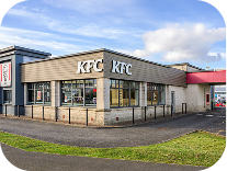 A KFC fast food restaurant with large windows, beige and gray exterior, and clear signage. The building is situated on a corner lot with a grassy area and a paved driveway.