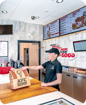 A KFC employee hands a large KFC Delivery bag to a customer across the counter inside a brightly lit KFC restaurant, with menu boards visible in the background.
