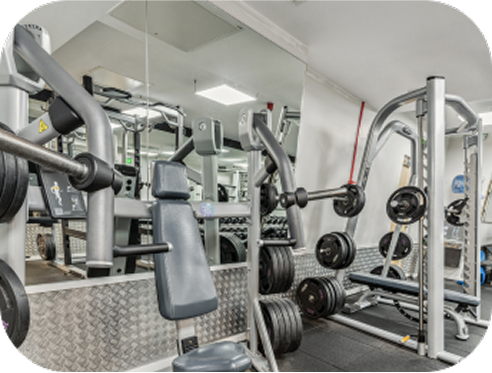 A gym with weightlifting equipment, including a bench press with weights, a weight machine, and a large mirror reflecting the workout area. The room is brightly lit with white walls and ceiling.