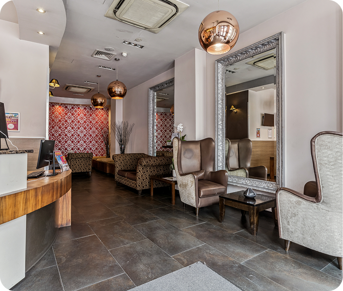 Modern hotel lobby with large mirrors, brown tile flooring, elegant armchairs, and copper ceiling lights. Patterned wallpaper adds color in the seating area, and a wooden reception desk is visible on the left.