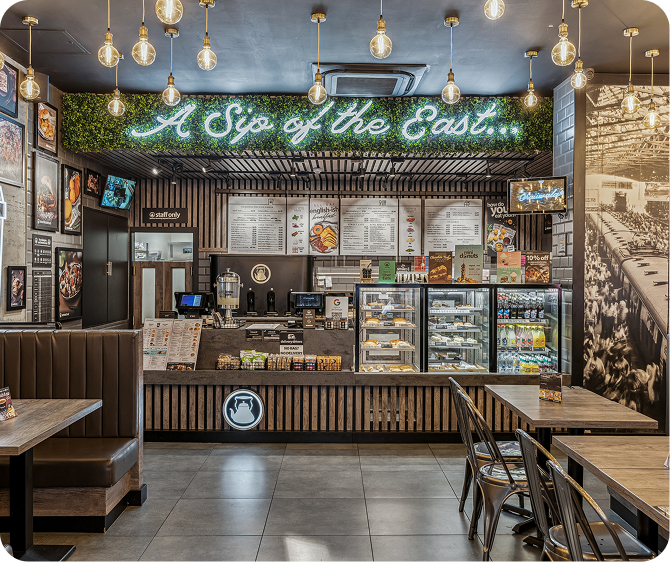 A stylish café with wooden décor, hanging lights, a neon sign reading A Sip of the East, menu boards above the counter, display cases with desserts and drinks, and booths and tables for seating.