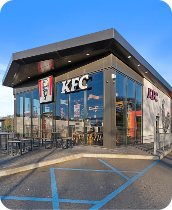 A modern KFC restaurant with large glass windows, outdoor seating, and visible KFC signage. There is a wheelchair-accessible parking space in the foreground under a clear blue sky.