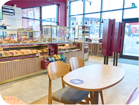 A brightly lit café with wooden tables and chairs, a display counter filled with pastries and snacks, large windows letting in natural light, and a view of the street outside.