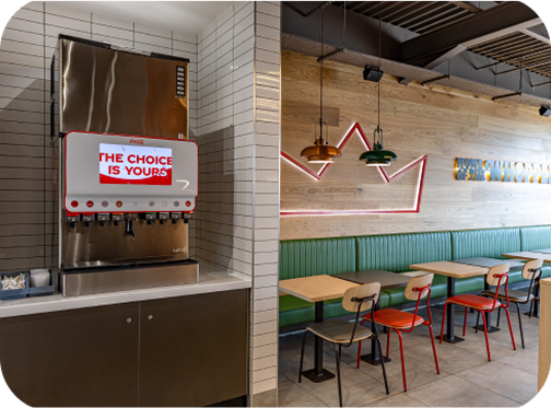 A modern fast-food restaurant interior with a drink dispenser on the left displaying THE CHOICE IS YOURS, and on the right, empty tables, colorful chairs, green booths, and stylish pendant lights.