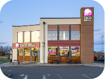 A modern Taco Bell restaurant with large windows, illuminated signs, and a parking lot in front. The sky is clear, suggesting it is either early morning or late evening.