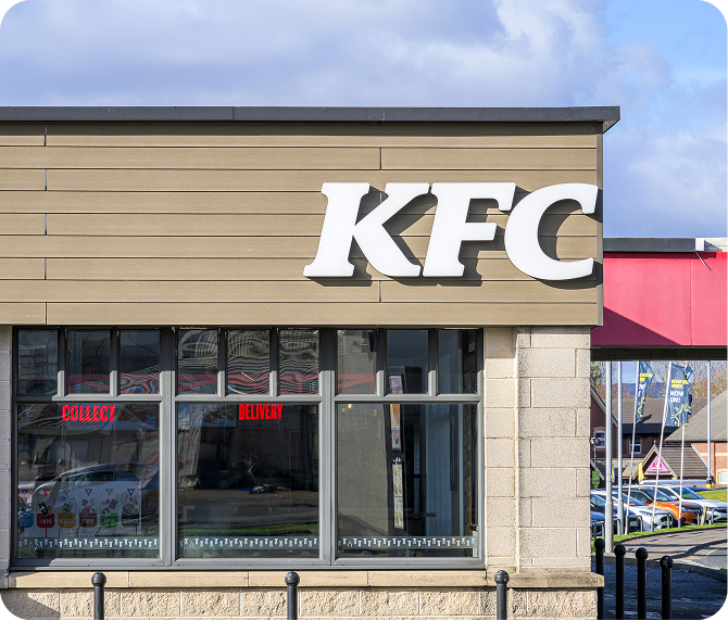 Exterior view of a KFC restaurant with large white KFC letters on the building; windows below display Collect and Delivery in red text. Cars and other buildings are visible in the background.