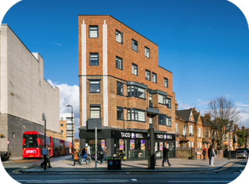 A modern brick building with a Taco Bell restaurant on the ground floor stands on a city street. People walk by on the sidewalk, and a red double-decker bus is visible on the left. The sky is clear and blue.