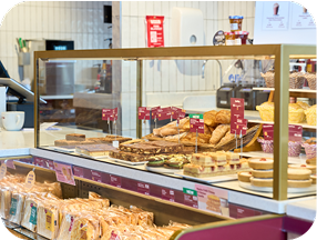 A bakery display case filled with various pastries, cakes, and sandwiches. Behind the glass, there are croissants, tarts, brownies, and other baked goods, with price tags visible. A coffee cup sits on the counter.