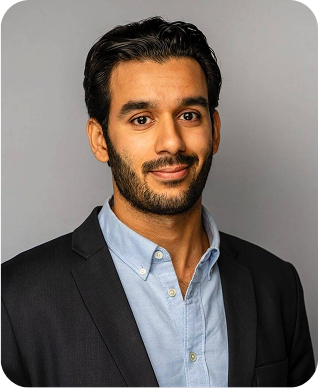 A man with short dark hair and a trimmed beard is wearing a dark blazer over a light blue shirt. He is standing against a plain gray background, looking at the camera with a slight smile.