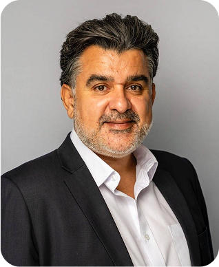 A middle-aged man with dark, wavy hair and a beard, wearing a dark suit jacket over a white dress shirt, poses against a plain light gray background.
